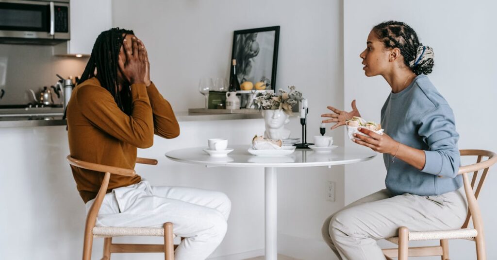 Side view of expressive Hispanic female in casual clothes arguing with African American boyfriend covering face with hands while sitting at table in kitchen at home