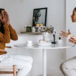 Side view of expressive Hispanic female in casual clothes arguing with African American boyfriend covering face with hands while sitting at table in kitchen at home